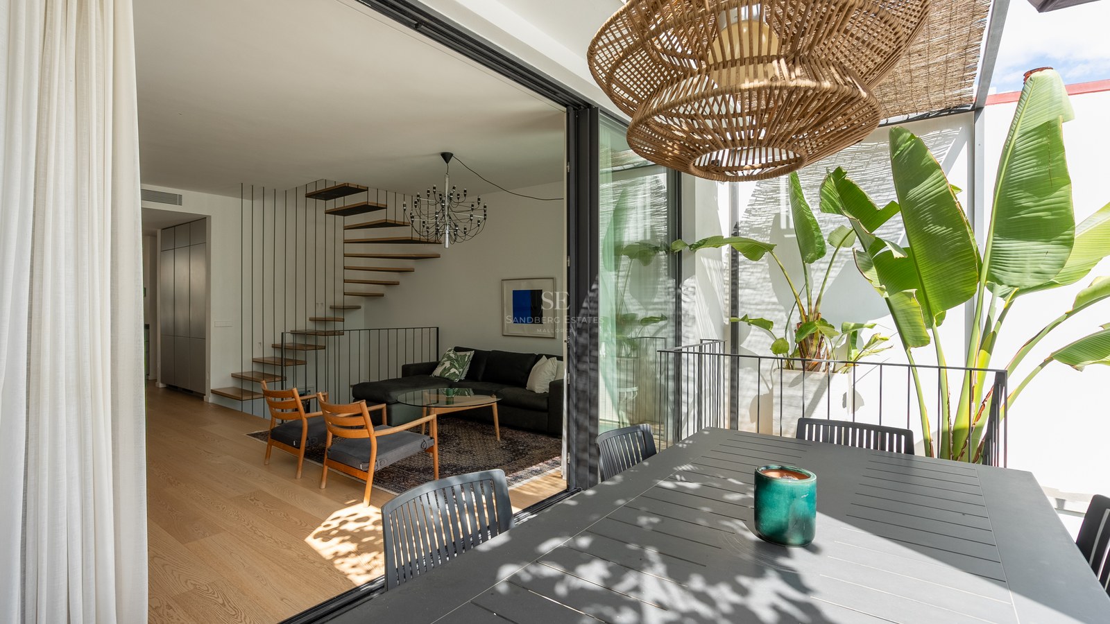 Salon moderne avec escalier en bois vu depuis une terrasse avec plantes tropicales à travers des baies vitrées.
