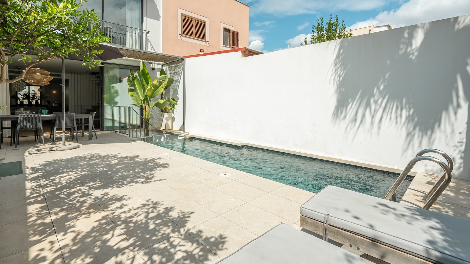 Une piscine étroite bleue sur une terrasse en dalles claires avec des chaises longues et un espace repas extérieur.