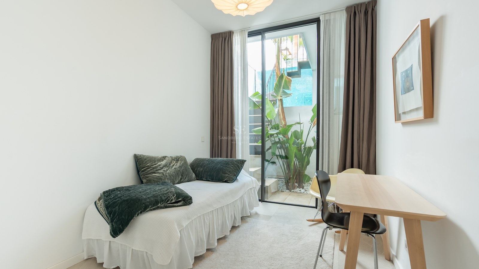 Chambre lumineuse avec un lit simple, un bureau en bois et une porte vitrée ouvrant sur un petit jardin extérieur.