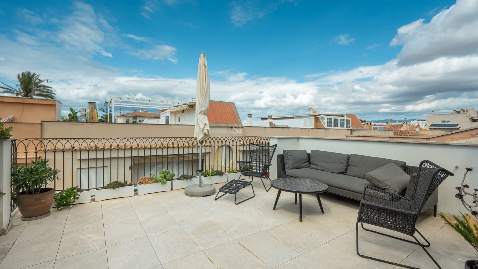 Terrasse de toit avec canapé gris, chaises noires et parasol blanc sous un ciel bleu.