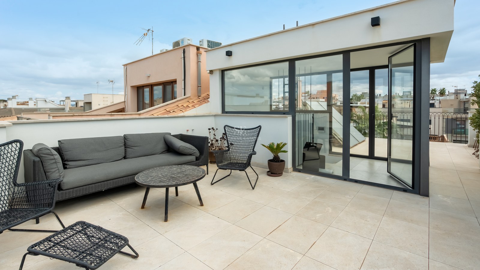 Terrasse sur le toit avec canapé gris, chaises design noires et grandes portes vitrées donnant sur la ville.