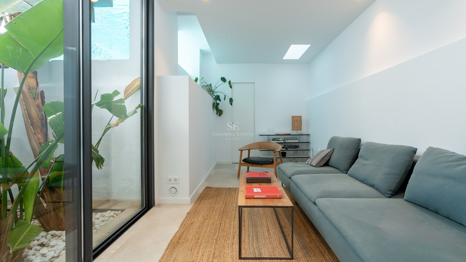 Salon moderne avec canapé bleu, table basse en bois, tapis en jute et baie vitrée donnant sur des plantes tropicales.