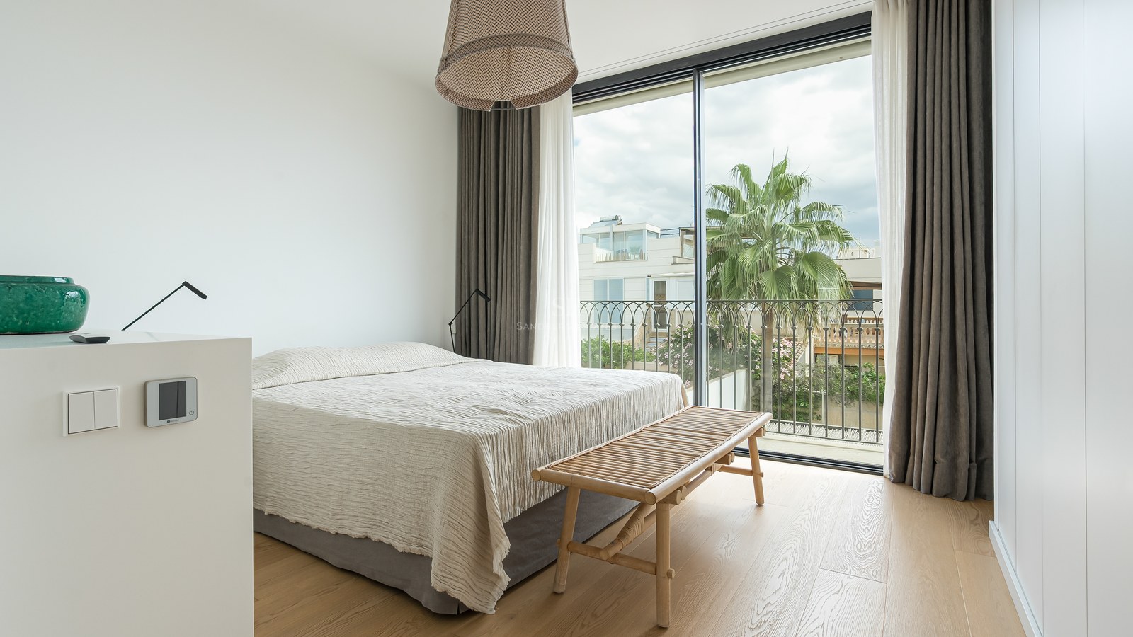 Chambre lumineuse avec baies vitrées, parquet en bois clair, grand lit et vue sur un palmier à l'extérieur.