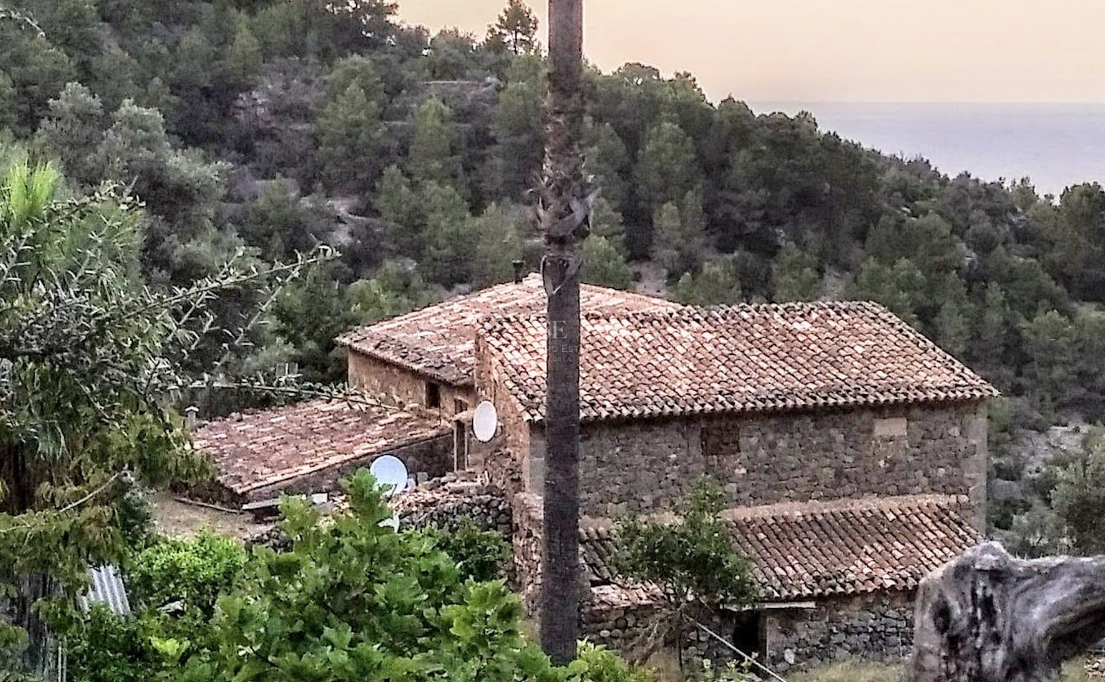 Traditional stone building with terracotta tiled roofs nestled in a lush green hillside overlooking the sea.