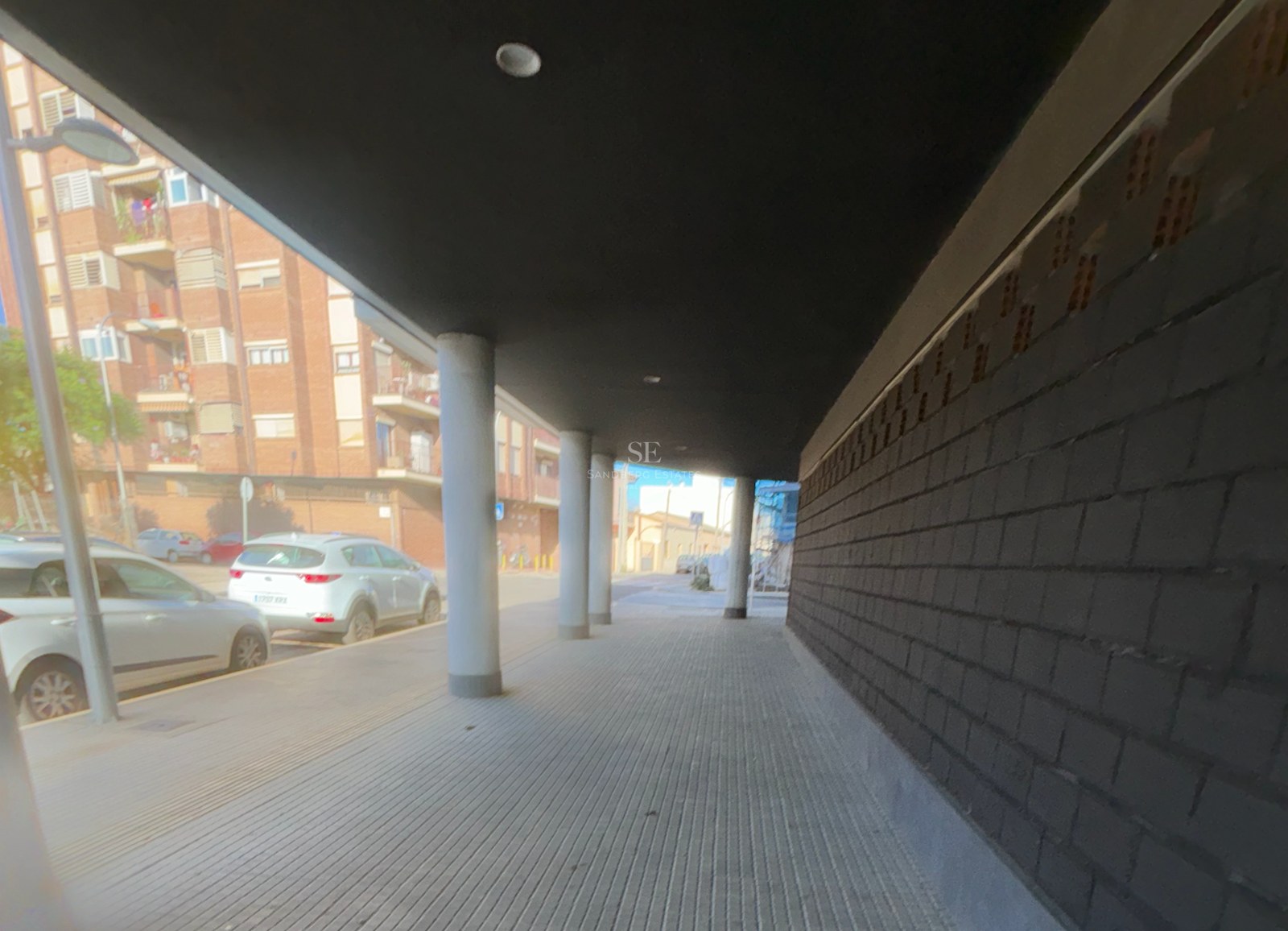 A covered sidewalk with concrete pillars and a dark brick wall next to a city street with parked cars.