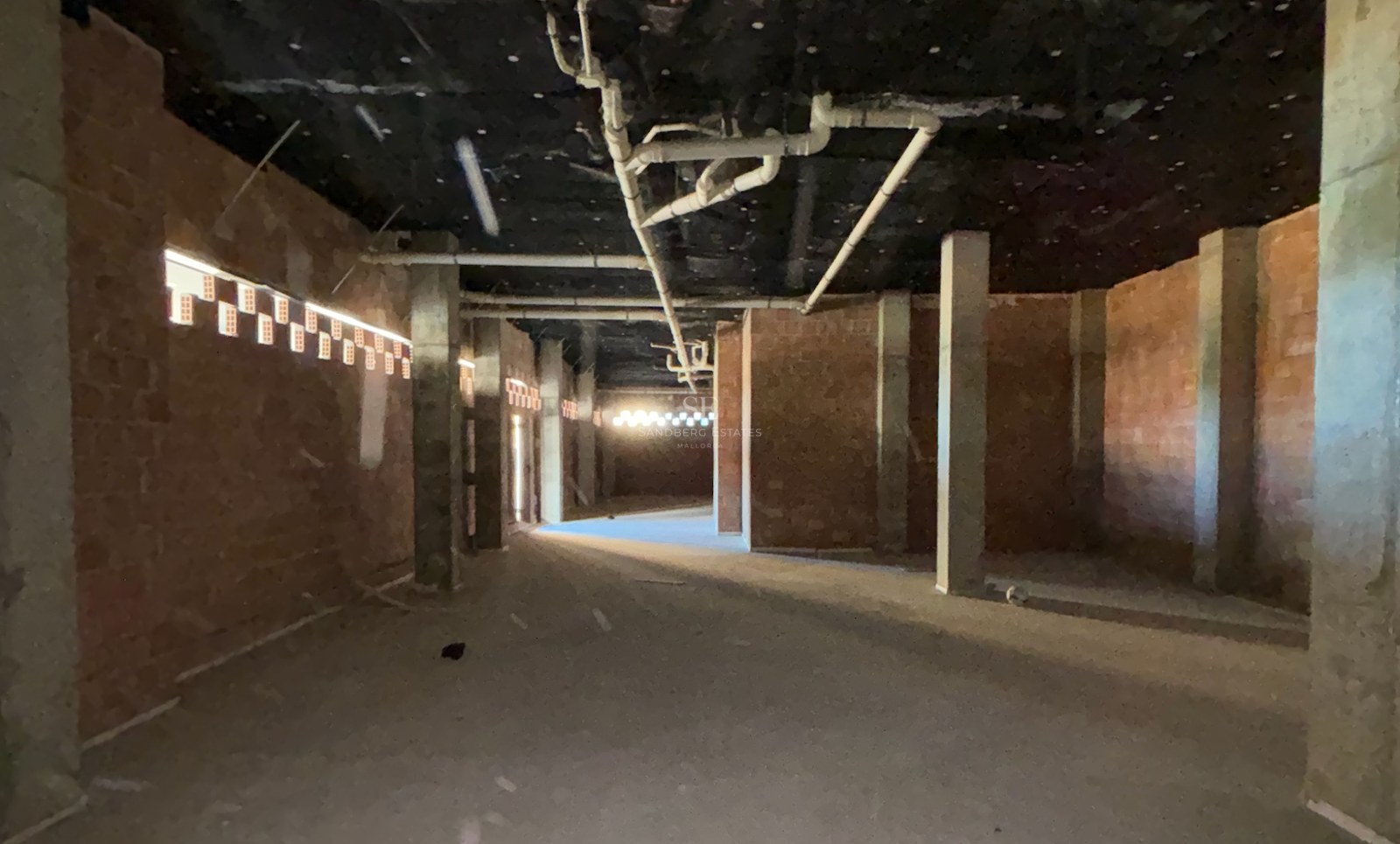 Raw interior space under construction with exposed brick walls, concrete pillars, and ceiling piping.