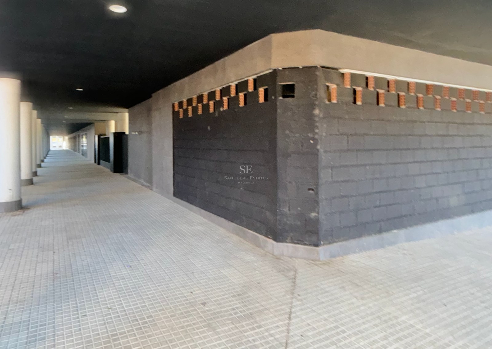 A long, covered outdoor walkway with dark brick walls, white columns, and grey paved flooring.