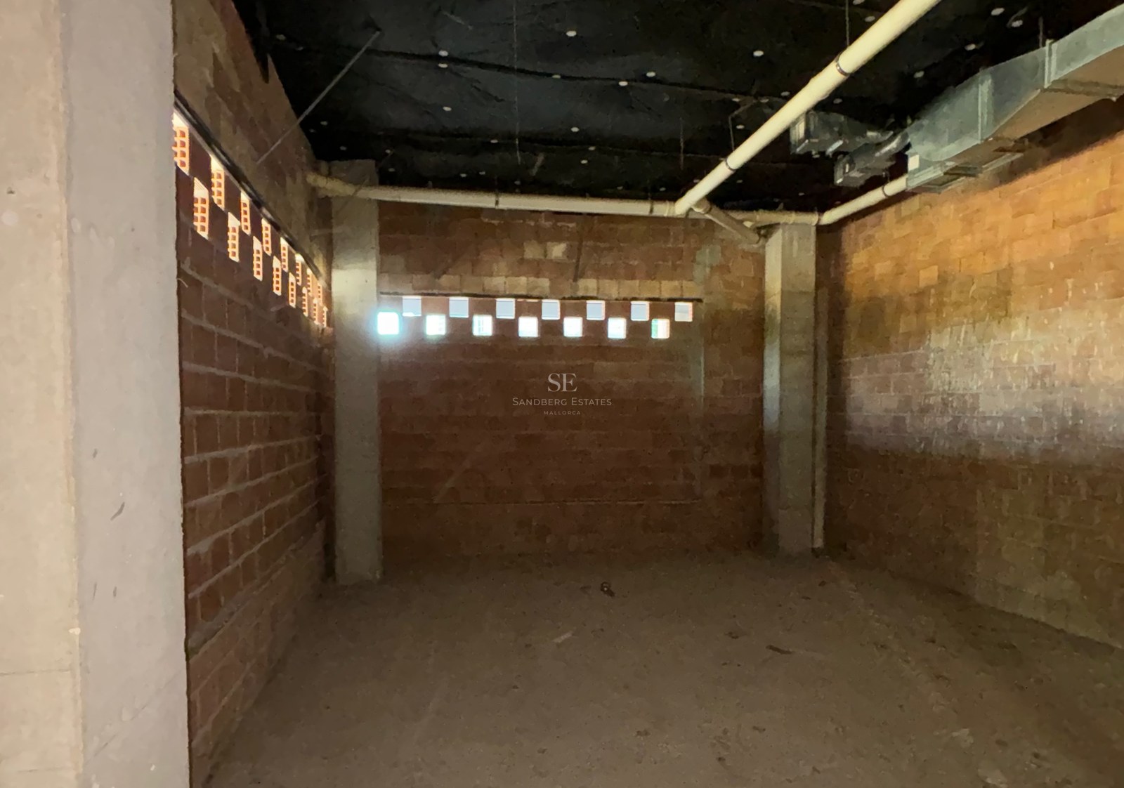Unfinished basement room with red brick walls, concrete pillars, and exposed utility pipes.