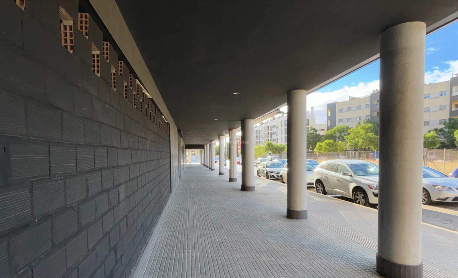Covered walkway featuring concrete pillars and a dark grey brick wall in an urban setting.