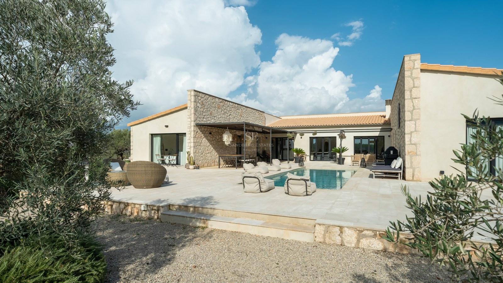 Extérieur d'une villa de luxe avec piscine centrale, murs en pierre naturelle et vaste terrasse pavée sous un ciel bleu.