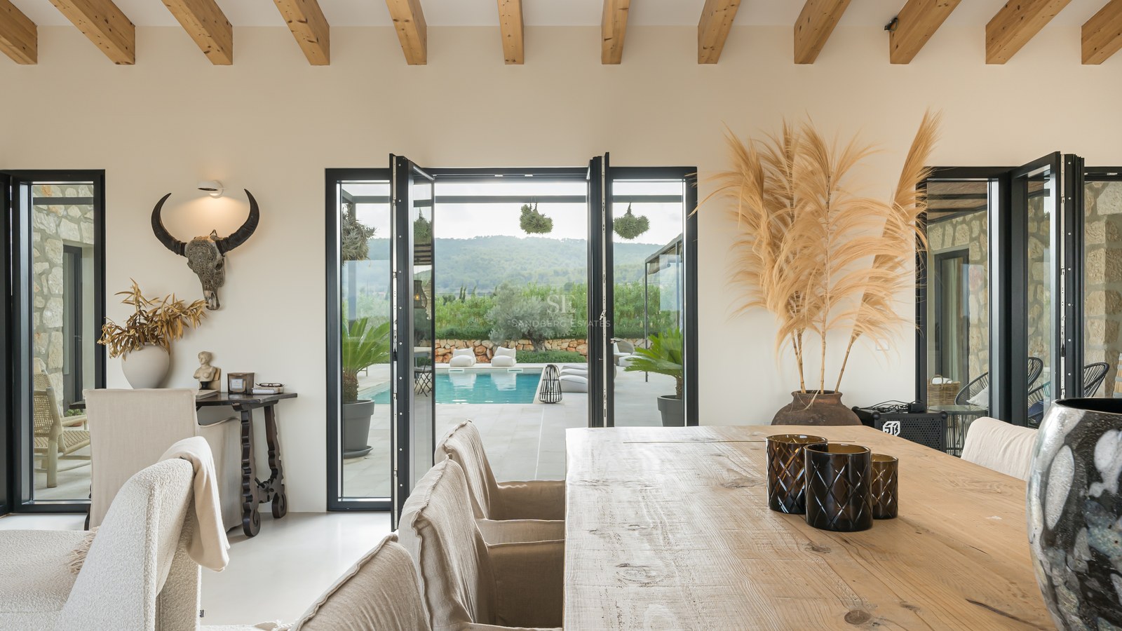 Salle à manger avec table en bois, poutres apparentes et larges baies vitrées donnant sur une terrasse et piscine.