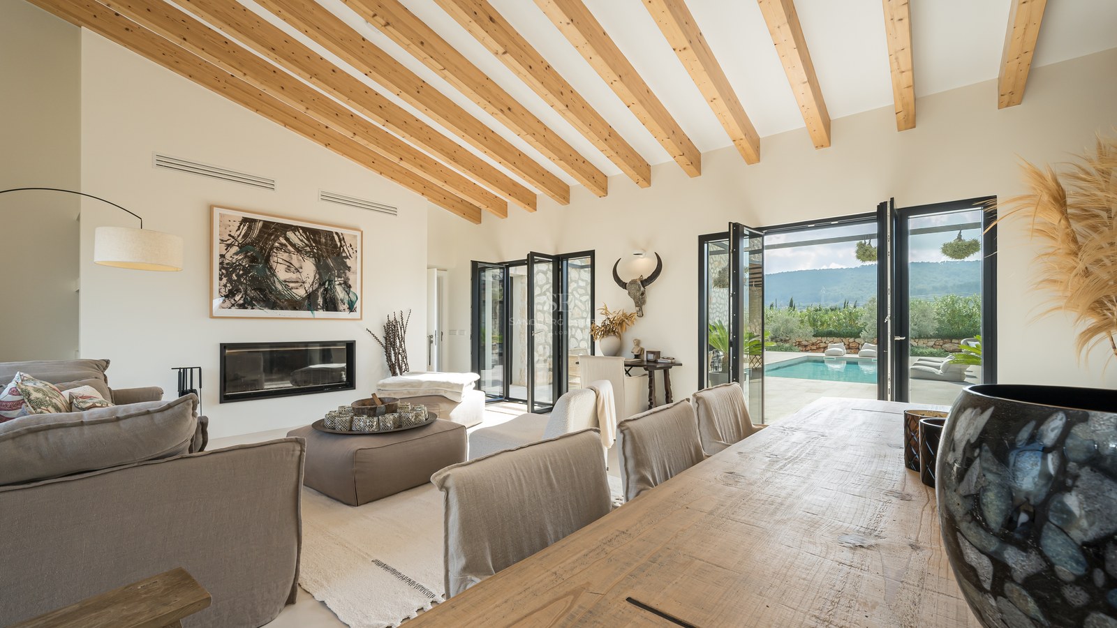 Salon élégant avec plafond voûté en bois, canapés en lin, cheminée et portes pliantes donnant sur la piscine.