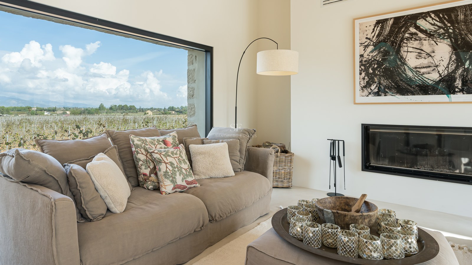 Salon moderne avec canapé taupe, cheminée et grande fenêtre donnant sur un vignoble sous un ciel nuageux.