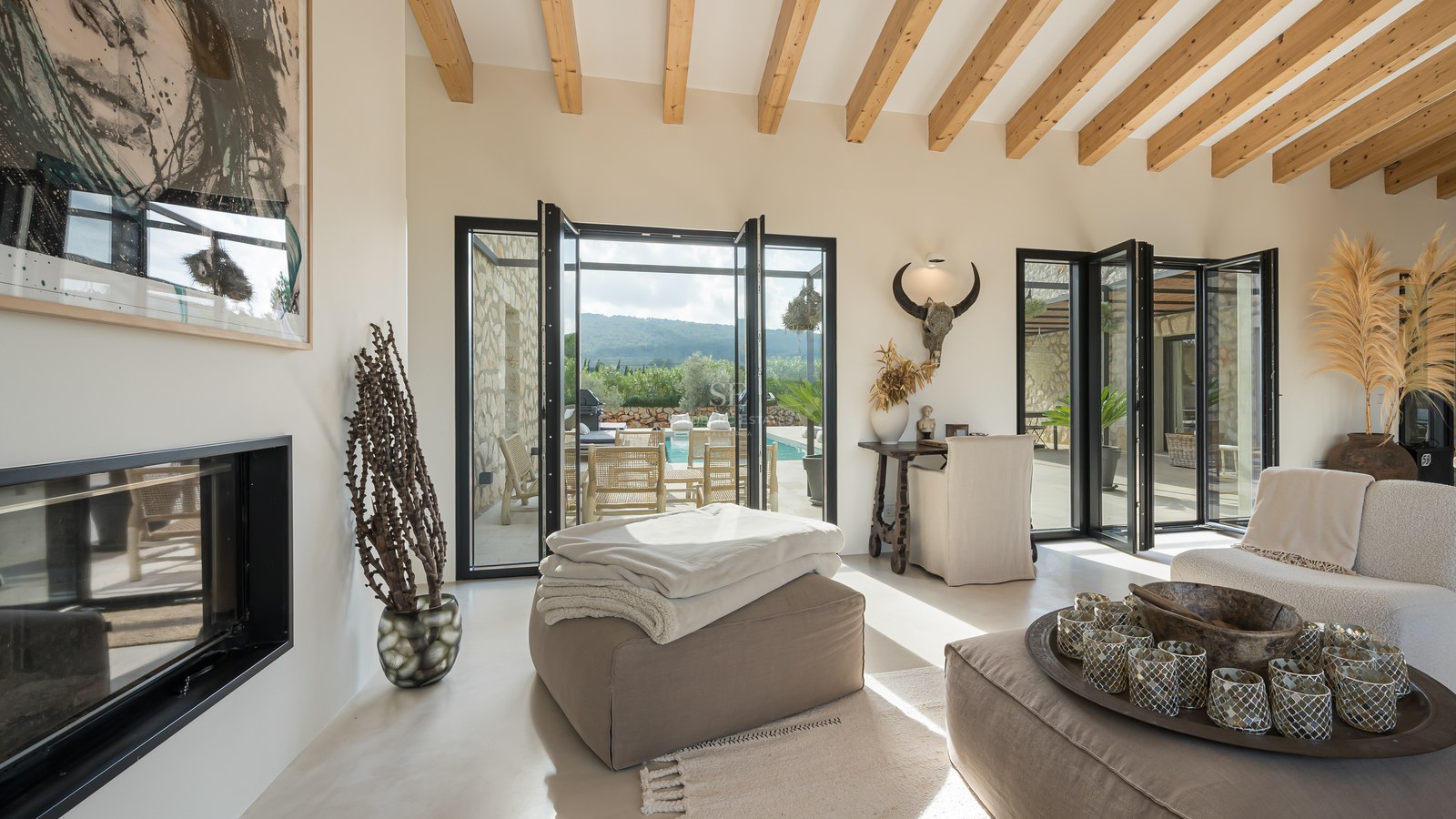 Salon moderne avec hauts plafonds à poutres, portes vitrées pliantes ouvrant sur une terrasse et une cheminée épurée.