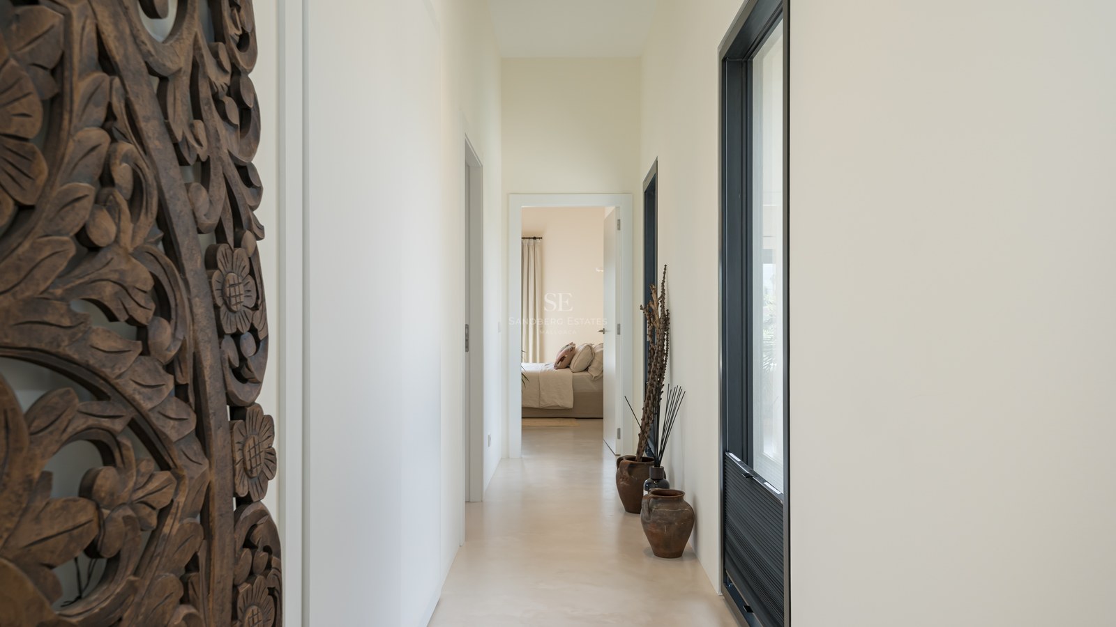 Couloir minimaliste avec sol en micro-ciment, panneau en bois sculpté et vases en terre cuite menant à une chambre.