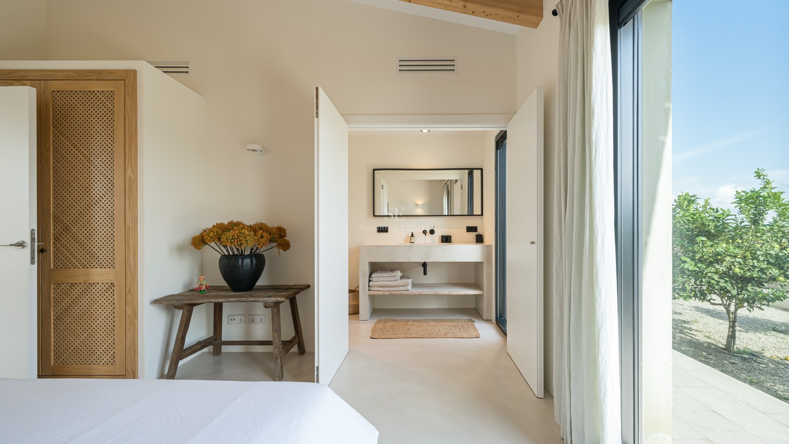 Chambre lumineuse avec poutres en bois et murs blancs s'ouvrant sur une salle de bains moderne en pierre.