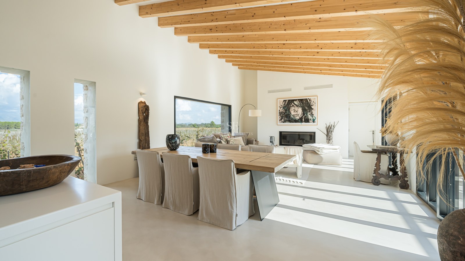Salle à manger lumineuse avec poutres en bois, grande table en chêne et baies vitrées donnant sur la campagne.