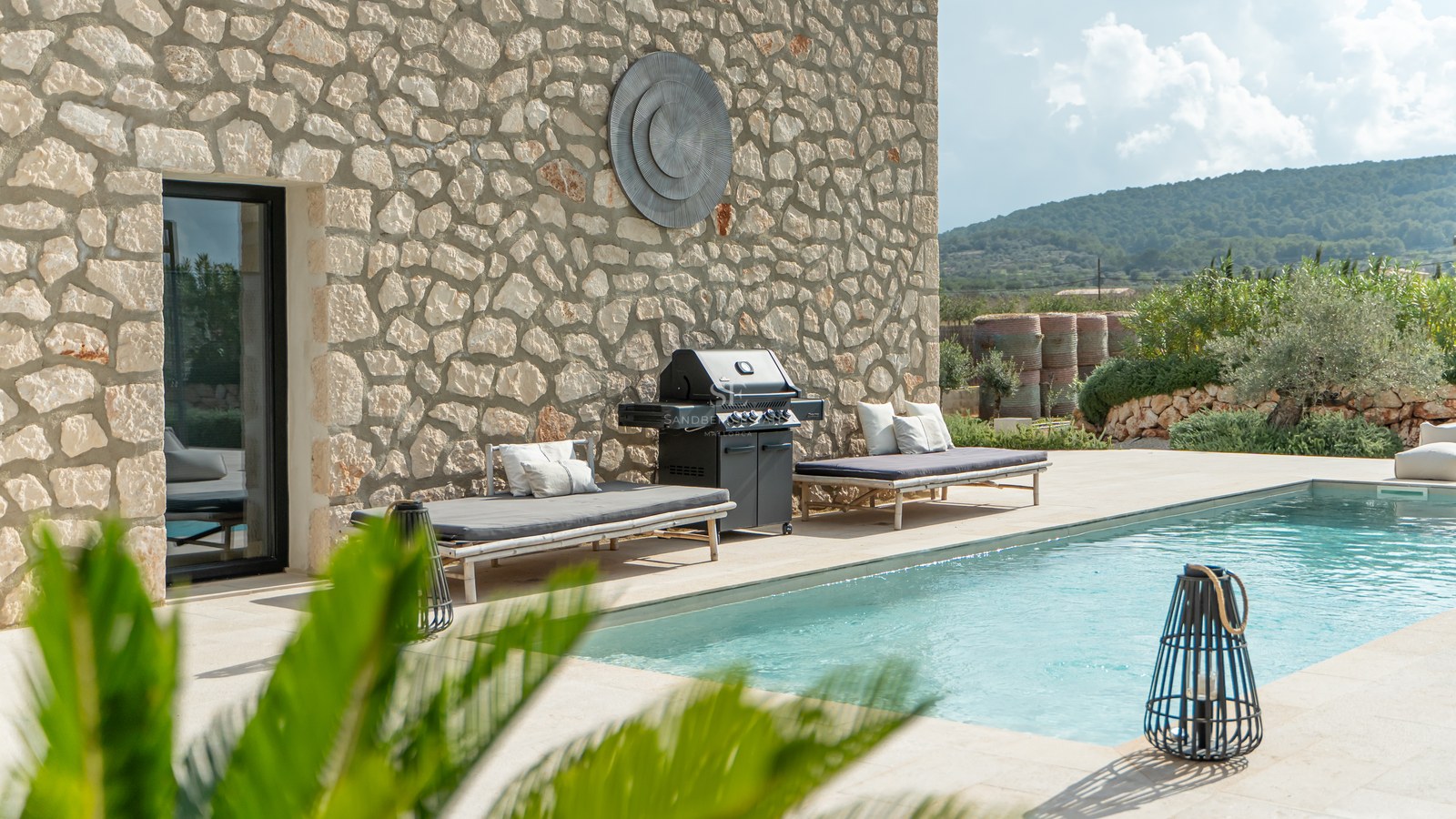 Piscine extérieure avec chaises longues, barbecue et mur en pierre naturelle sous un ciel azur.