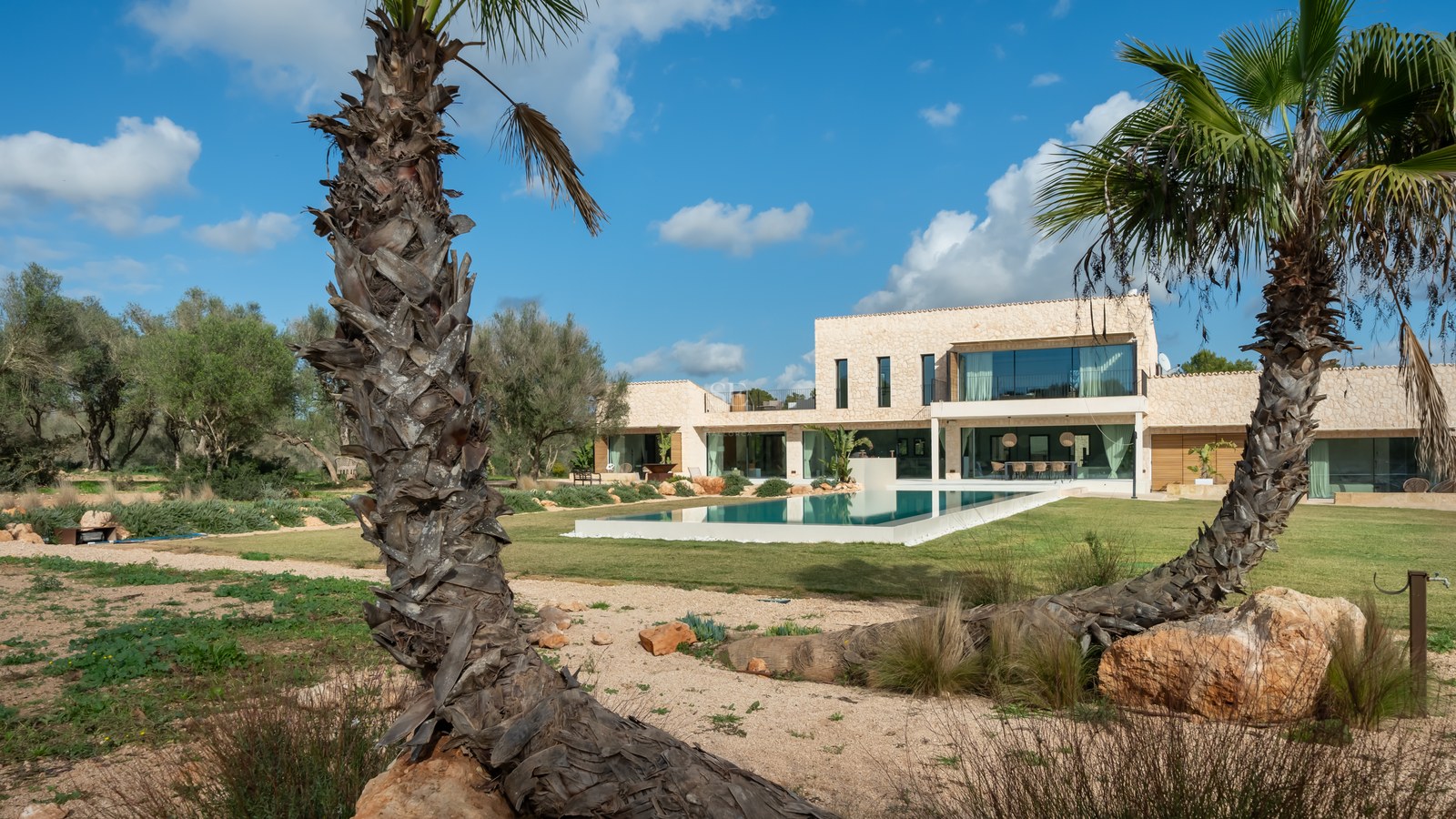 Villa moderne de deux étages avec une grande piscine rectangulaire, pelouse et palmiers sous un ciel bleu.