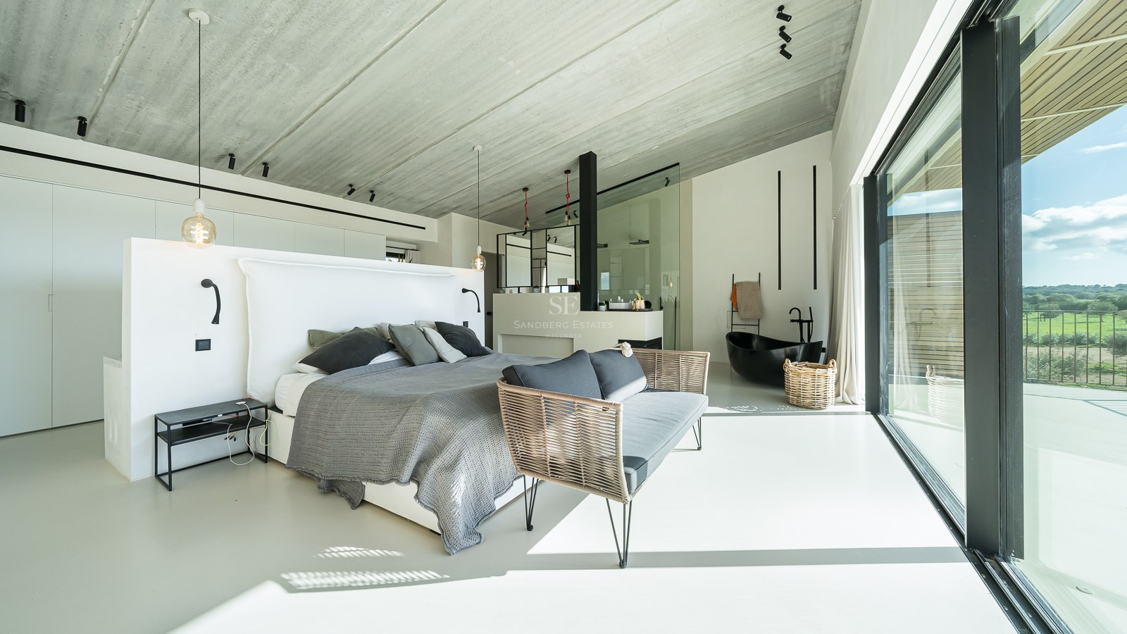 Chambre principale moderne avec lit gris, plafond en béton et baignoire îlot noire intégrée.
