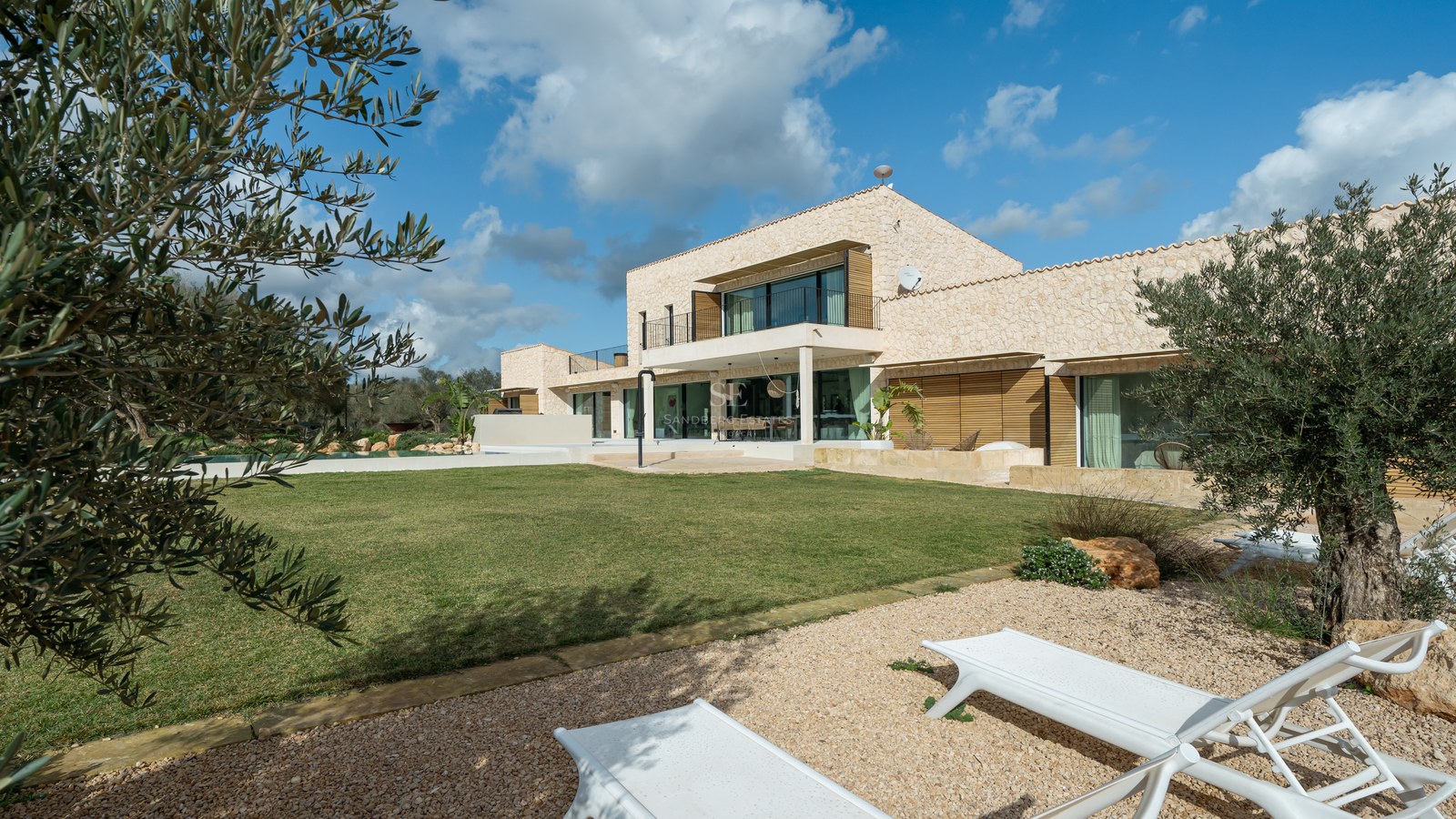 Villa contemporaine de deux étages avec murs en pierre, grandes baies vitrées et jardin sous un ciel bleu.