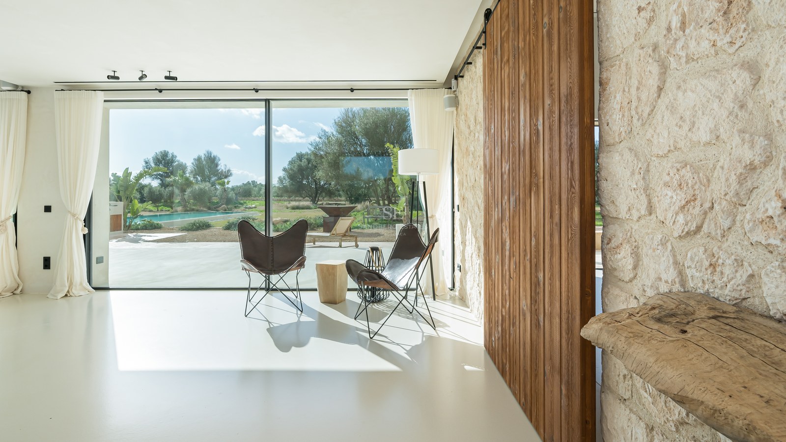 Salon lumineux avec baies vitrées, fauteuils butterfly, porte coulissante en bois et mur en pierre naturelle.