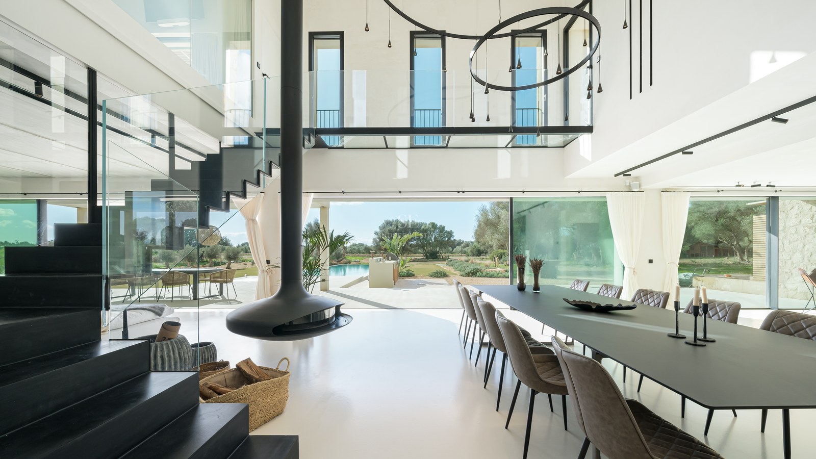 Salle à manger lumineuse à double hauteur avec cheminée suspendue noire, parois vitrées et vue sur le jardin et la piscine.