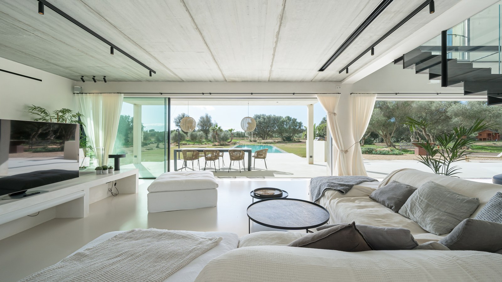 Intérieur de salon lumineux avec canapés blancs, ouvrant sur une terrasse, un coin repas extérieur et une piscine.