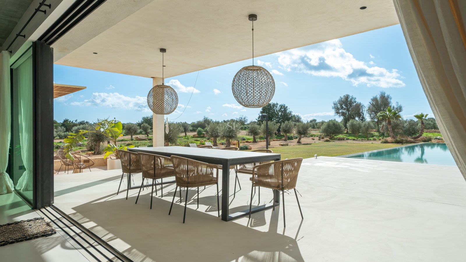 Espace repas extérieur couvert avec table moderne et chaises en rotin surplombant un jardin d'oliviers.