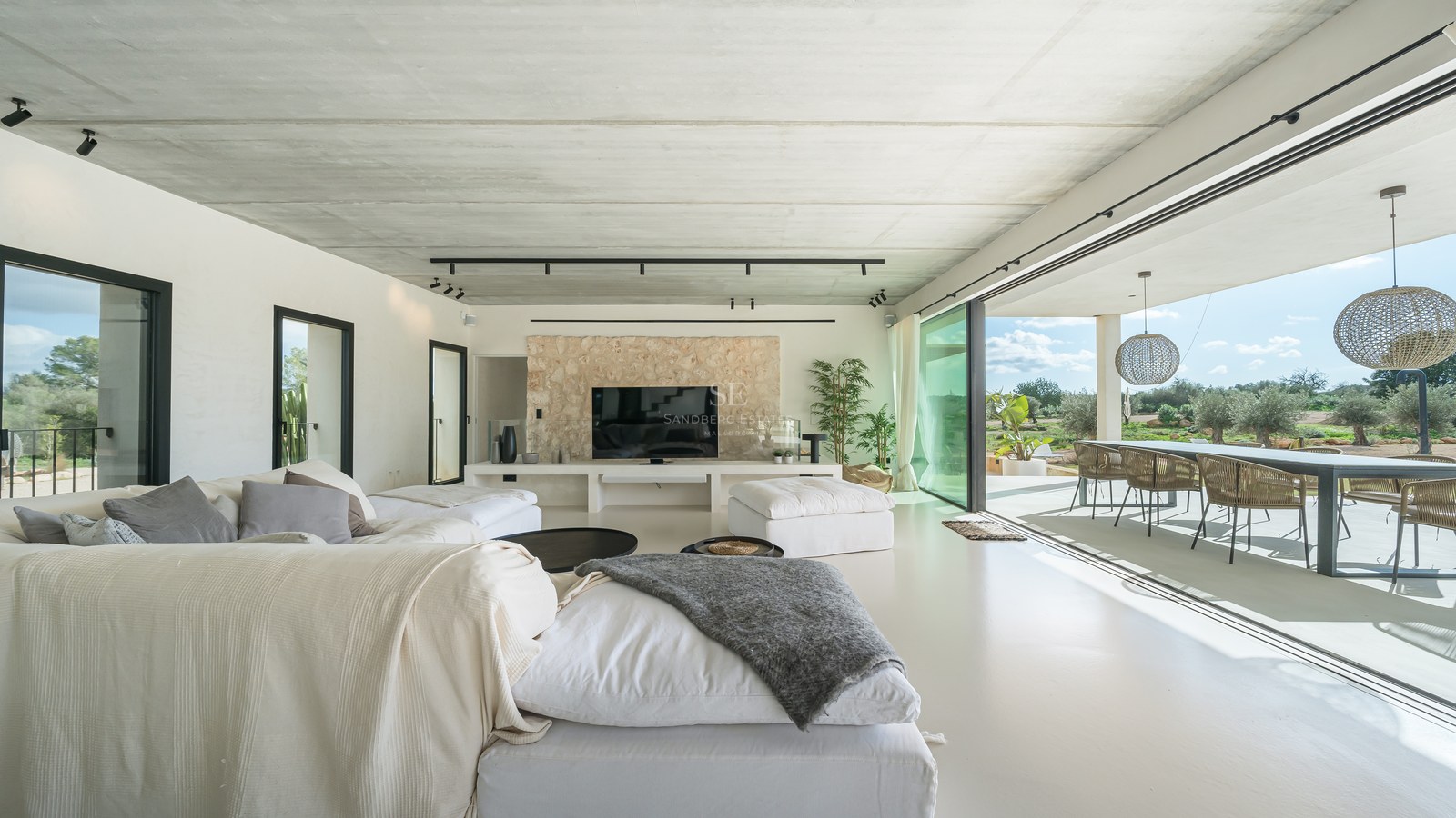 Salon moderne avec canapés blancs, plafond en béton, mur en pierre et baies vitrées ouvrant sur une terrasse.