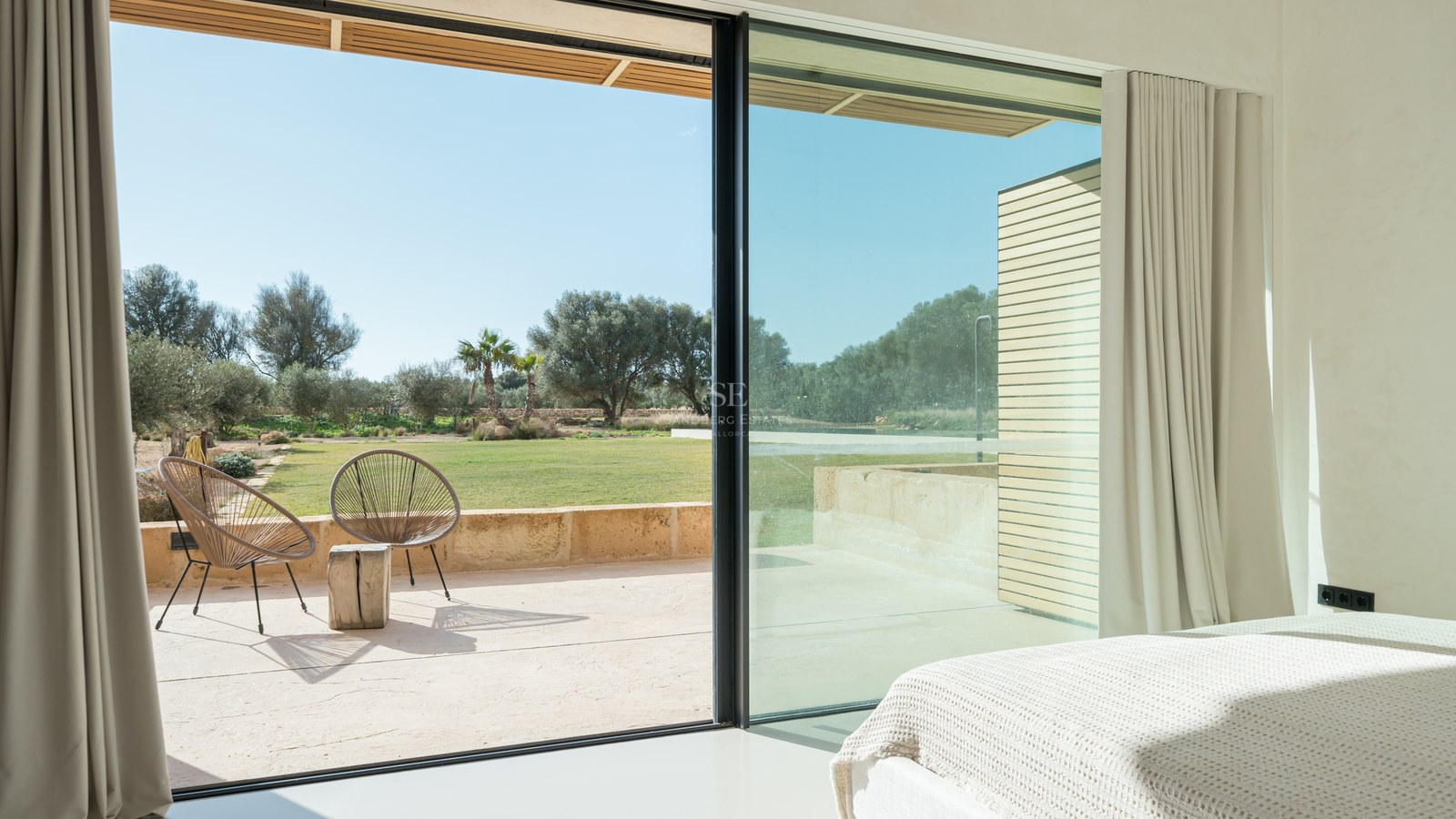 Vue depuis une chambre à travers des baies vitrées coulissantes sur une terrasse ensoleillée et un jardin paysager.