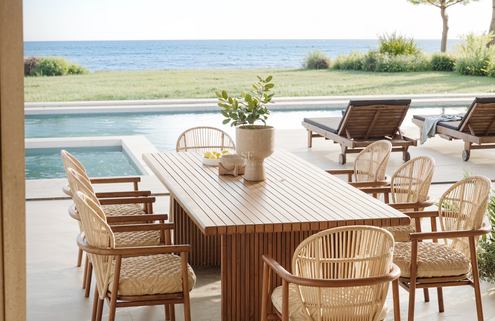 Table à manger en bois et chaises en osier sur une terrasse surplombant une piscine et la mer Méditerranée.