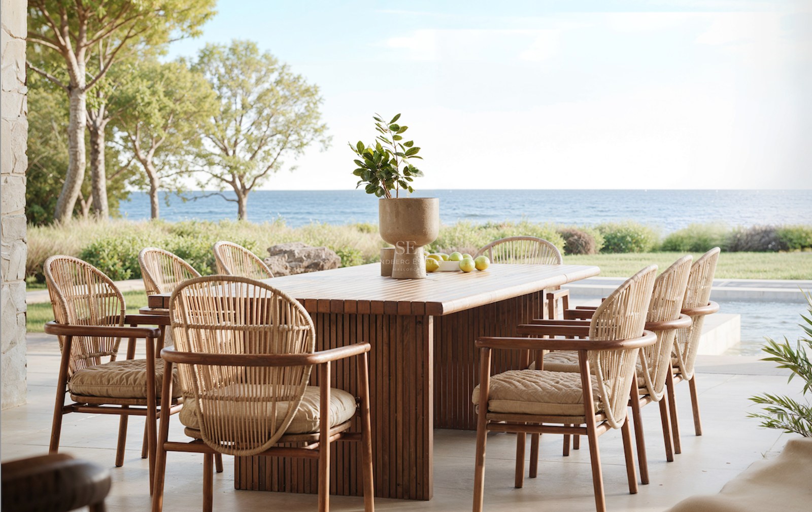 Une table à manger en bois et six chaises en osier sur une terrasse en pierre surplombant la mer Méditerranée.