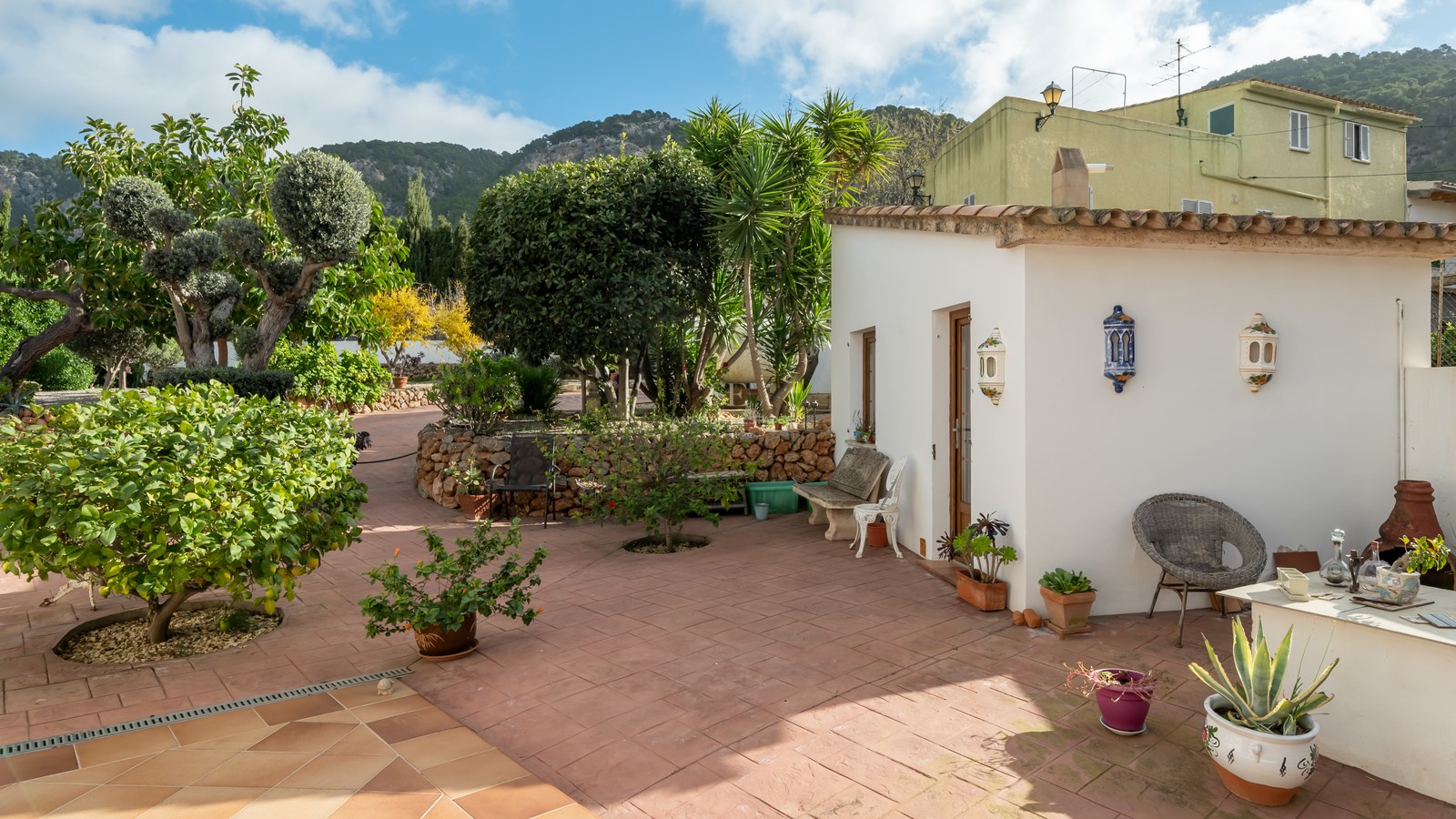 Sun-drenched terracotta patio featuring a whitewashed annex, lush Mediterranean gardens, and mountain views.