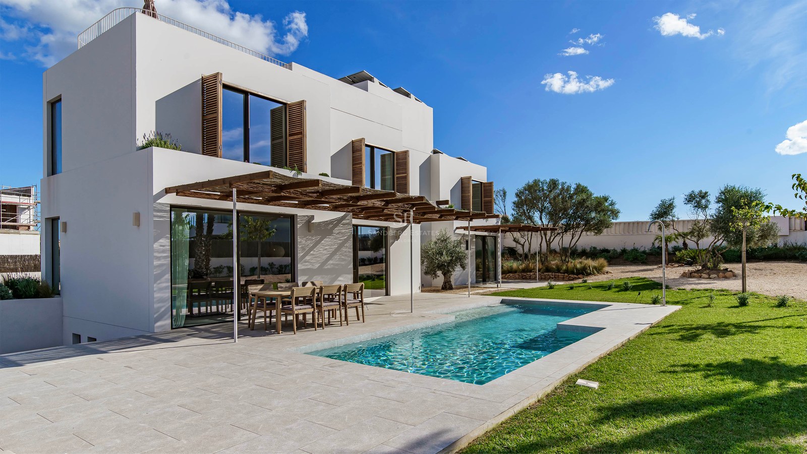 Exteriör av en vit modern villa med pool, träpergola och gräsmatta under en blå himmel.
