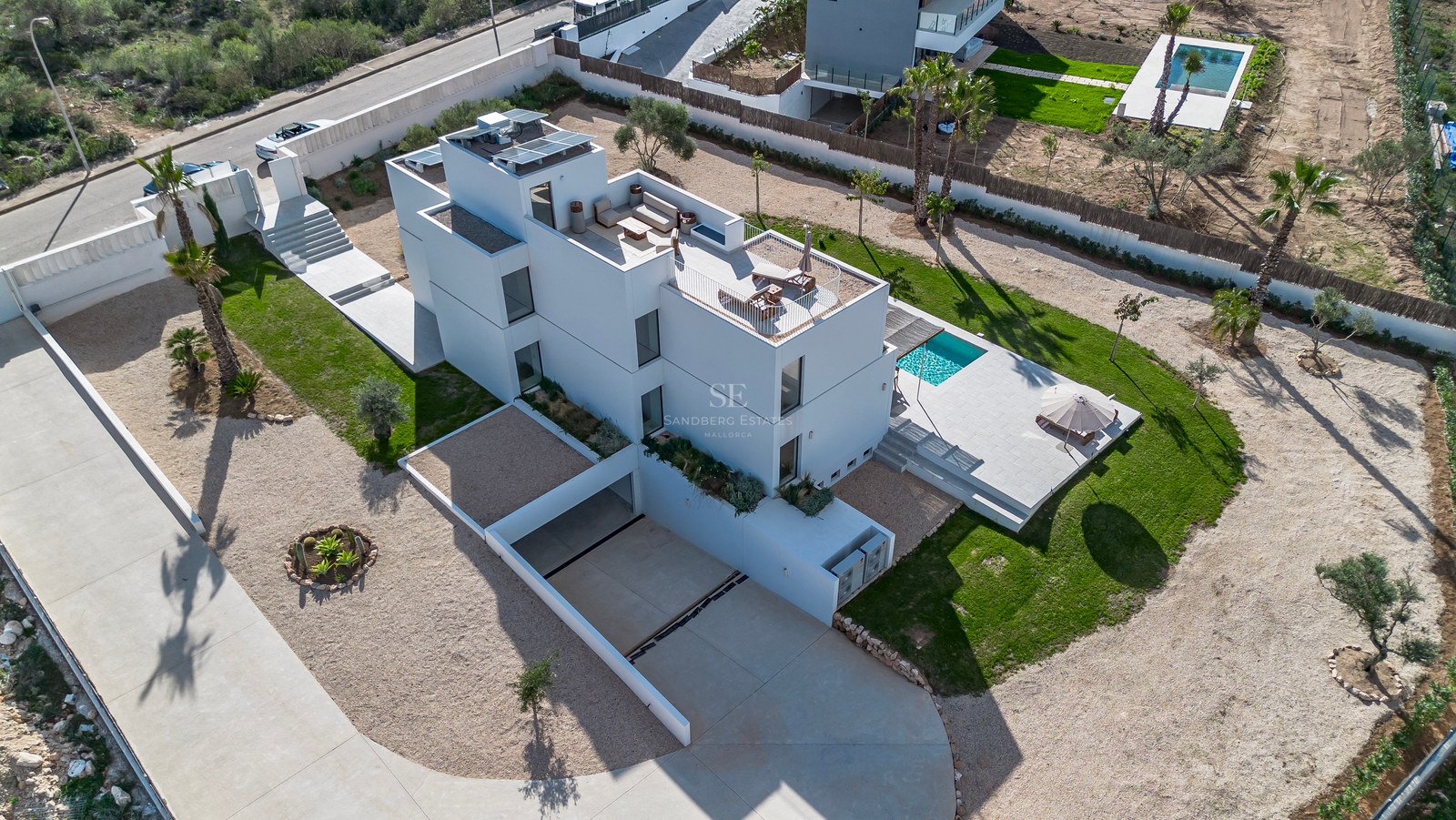 Flygfoto över en vit modern villa i tre plan med pool, anlagd trädgård och solpaneler.