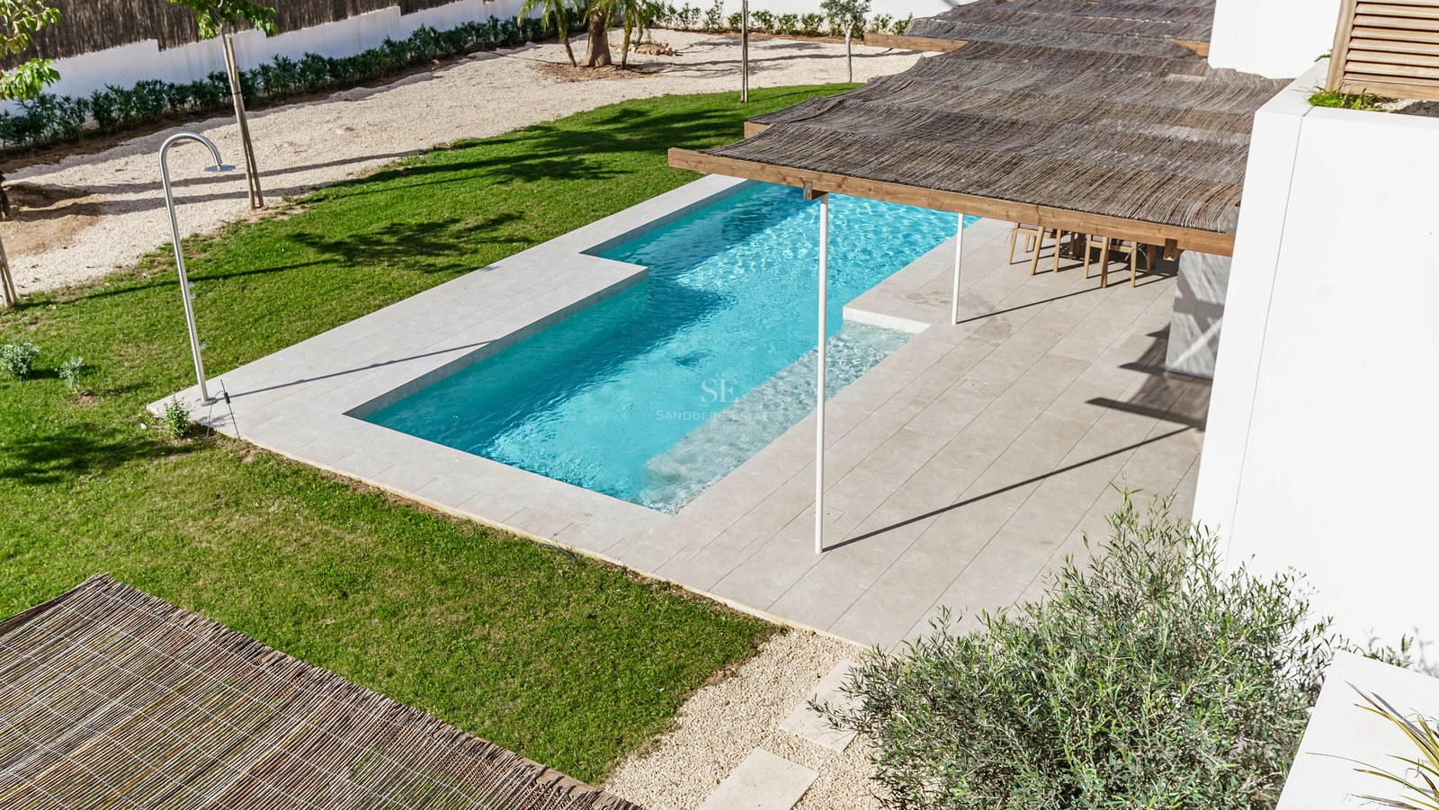 Flygvy över en turkos rektangulär pool omgiven av stenterrass, gräsmatta och en pergola med vasstak.