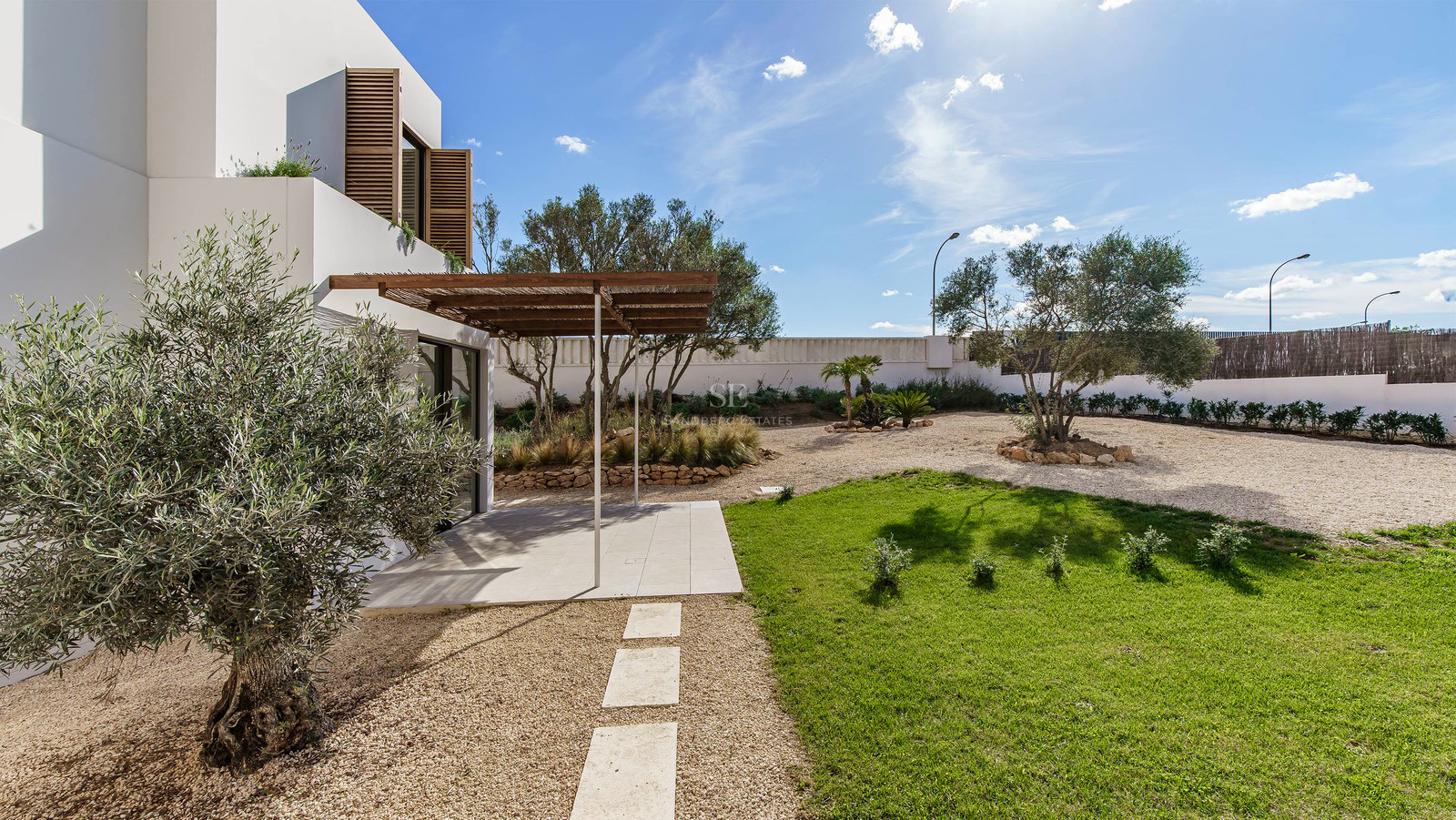 Exteriör av modern villa med olivträdgård, gräsmatta, grusgångar och träpergola under en klarblå himmel.