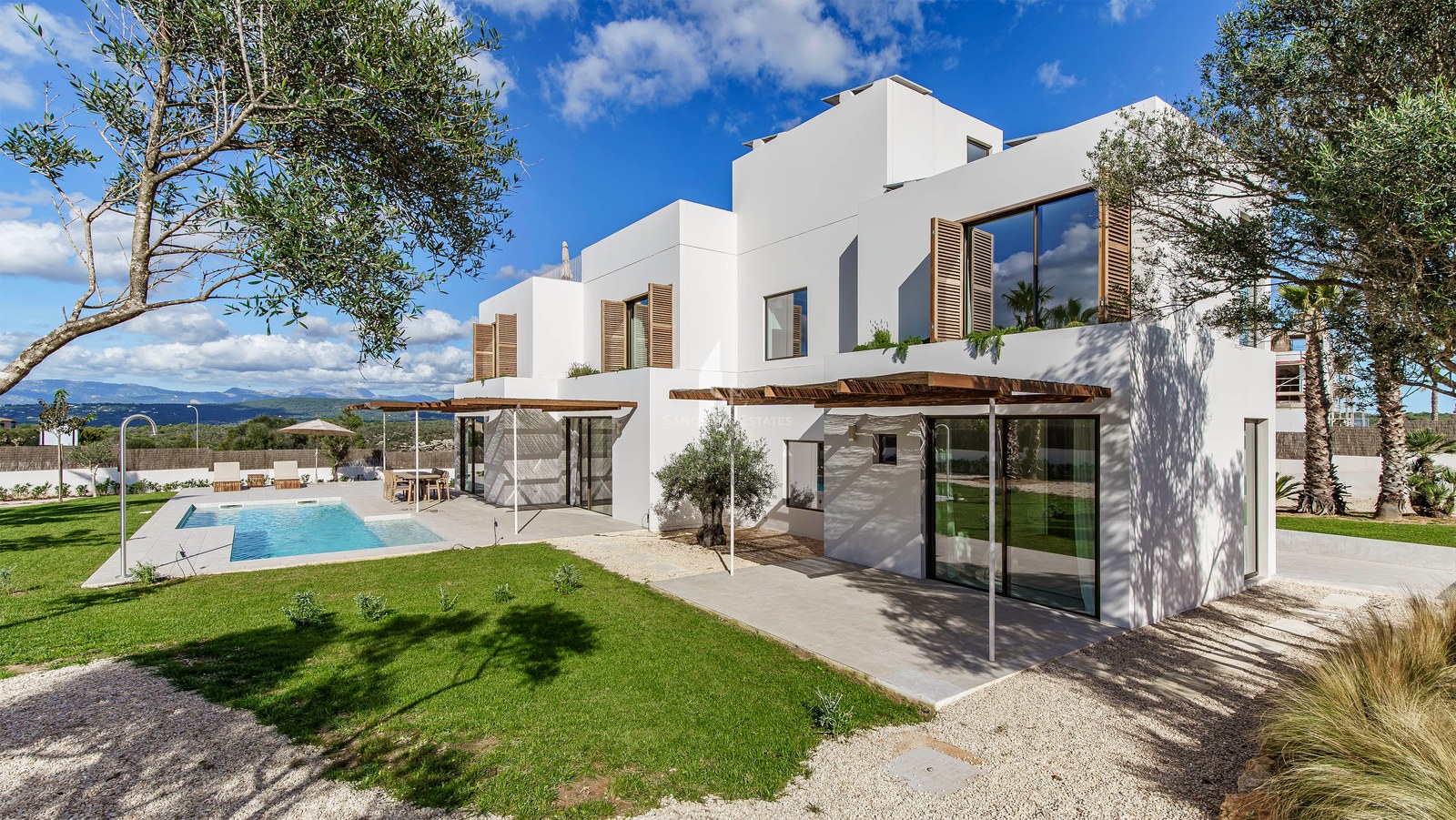Modern vit villa med turkos pool, träpergola och olivträd under en blå himmel.
