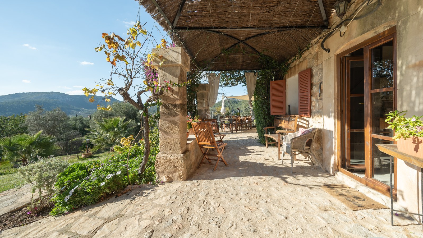 Terrasse en pierre rustique avec toit de chaume, meubles en bois et vue sur les collines verdoyantes.