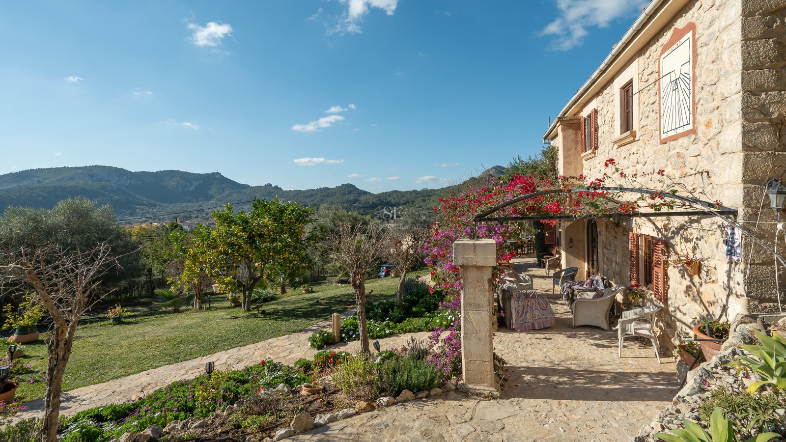 Terrasse en pierre avec mobilier extérieur, bougainvilliers roses et vue sur les montagnes sous un ciel bleu.