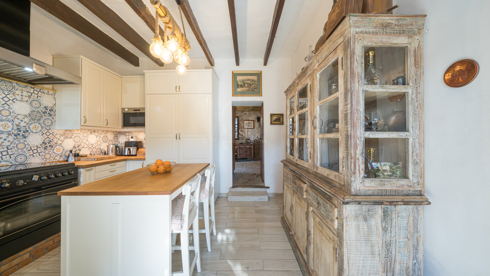 Cuisine lumineuse avec poutres de plafond en bois sombre, armoires blanches, îlot avec tabourets et grand buffet vintage.