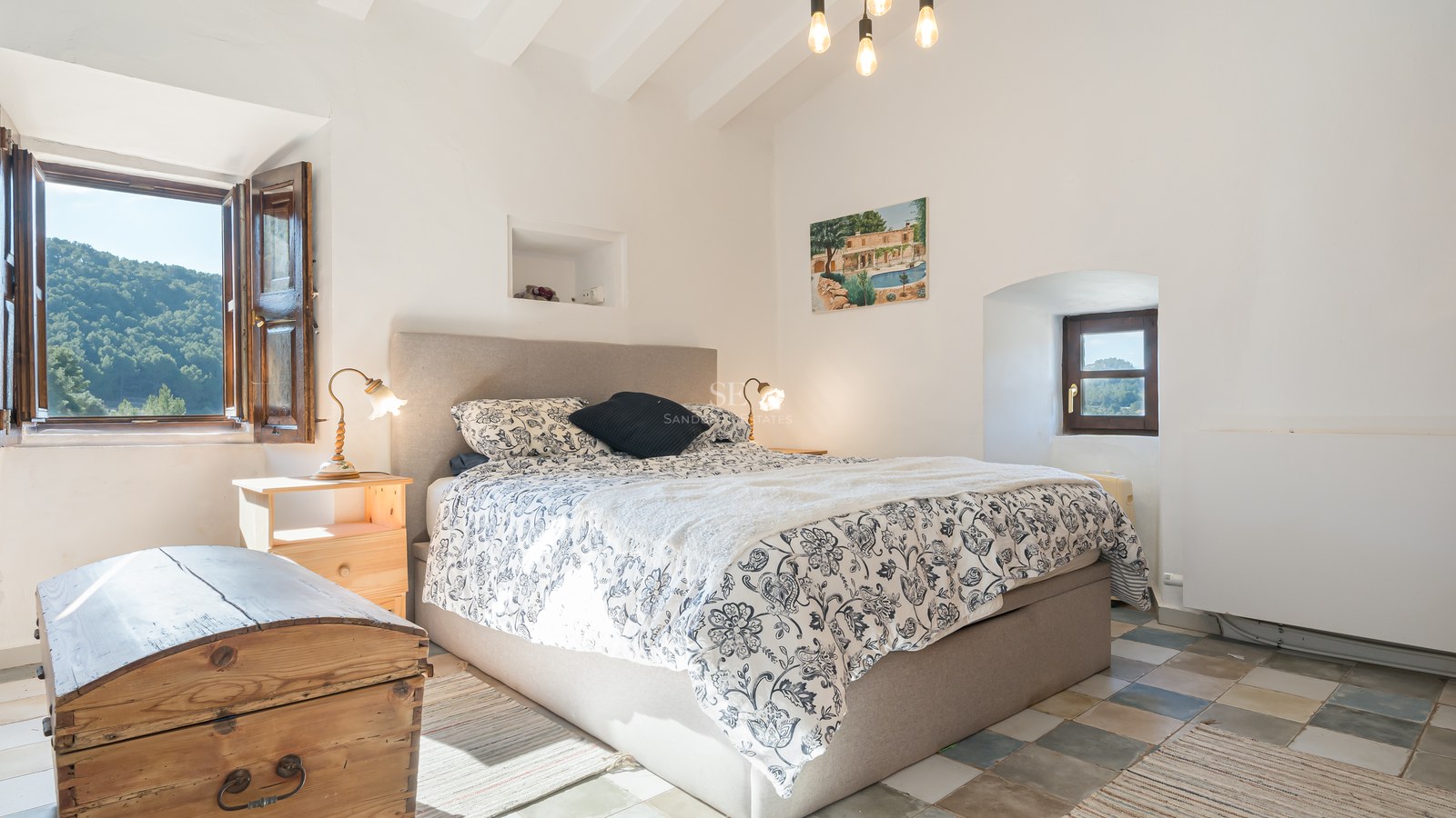 Chambre lumineuse avec poutres blanches, lit rembourré, linge de lit floral et tables de chevet en bois rustique.