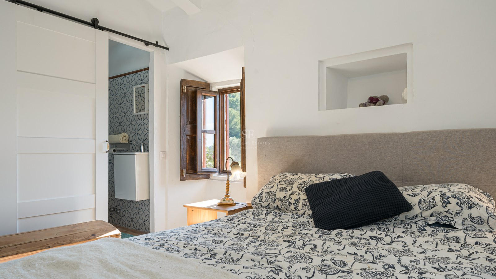 Chambre ensoleillée avec couvre-lit à motifs, volet en bois traditionnel et vue sur une salle de bain carrelée.