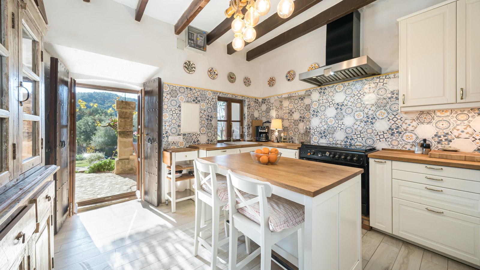 Cuisine rustique avec poutres en bois, carreaux artisanaux, îlot central avec tabourets et vue sur cour.
