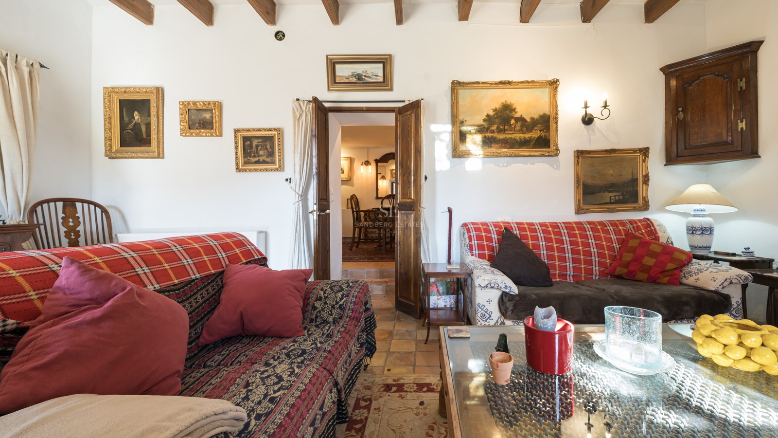 Salon de style rustique avec sol en terre cuite, poutres en bois sombre et tableaux classiques sur murs blancs.