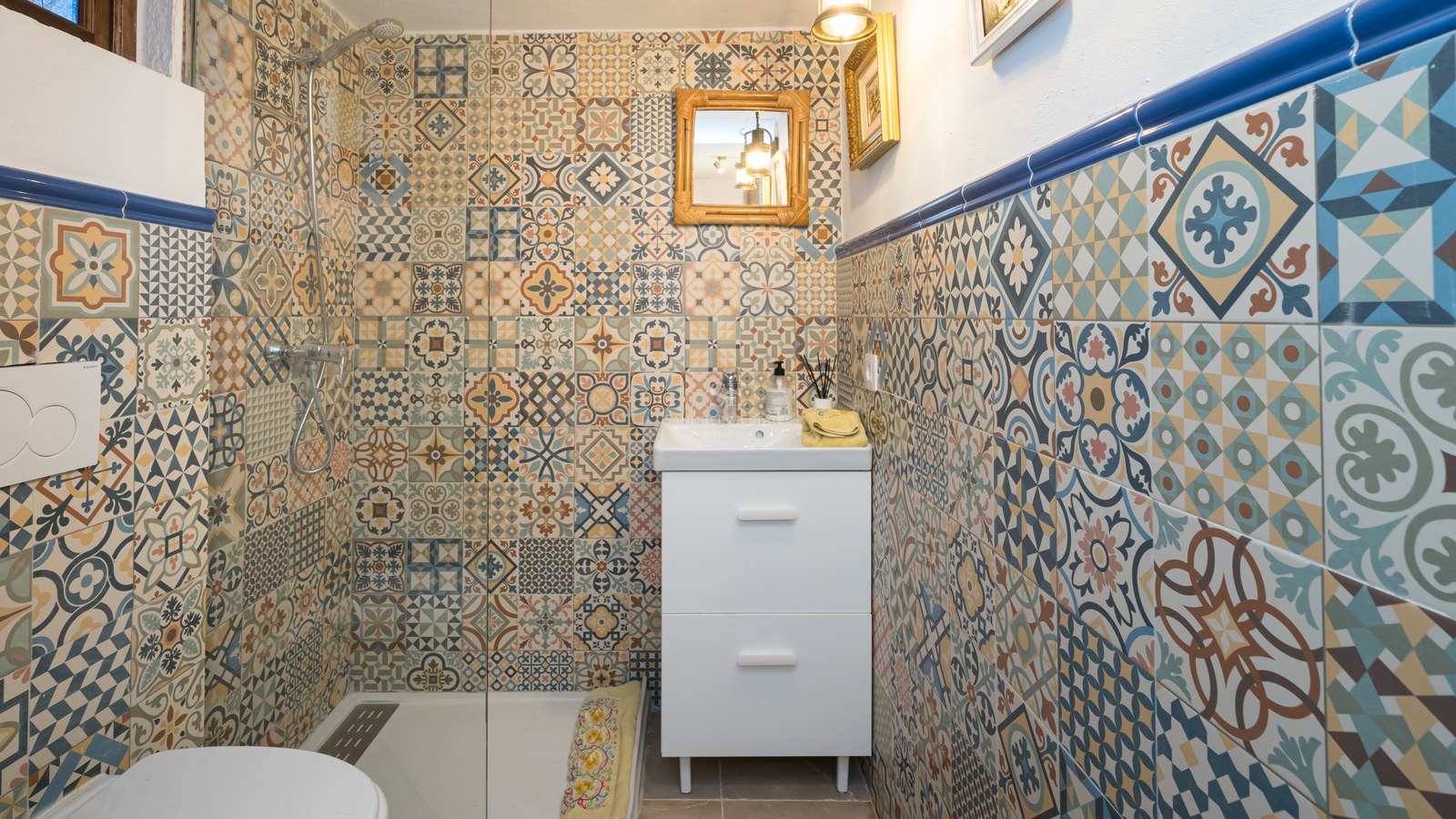 Salle de bain avec carrelage à motifs géométriques bleus et ocres, meuble vasque blanc et douche vitrée.