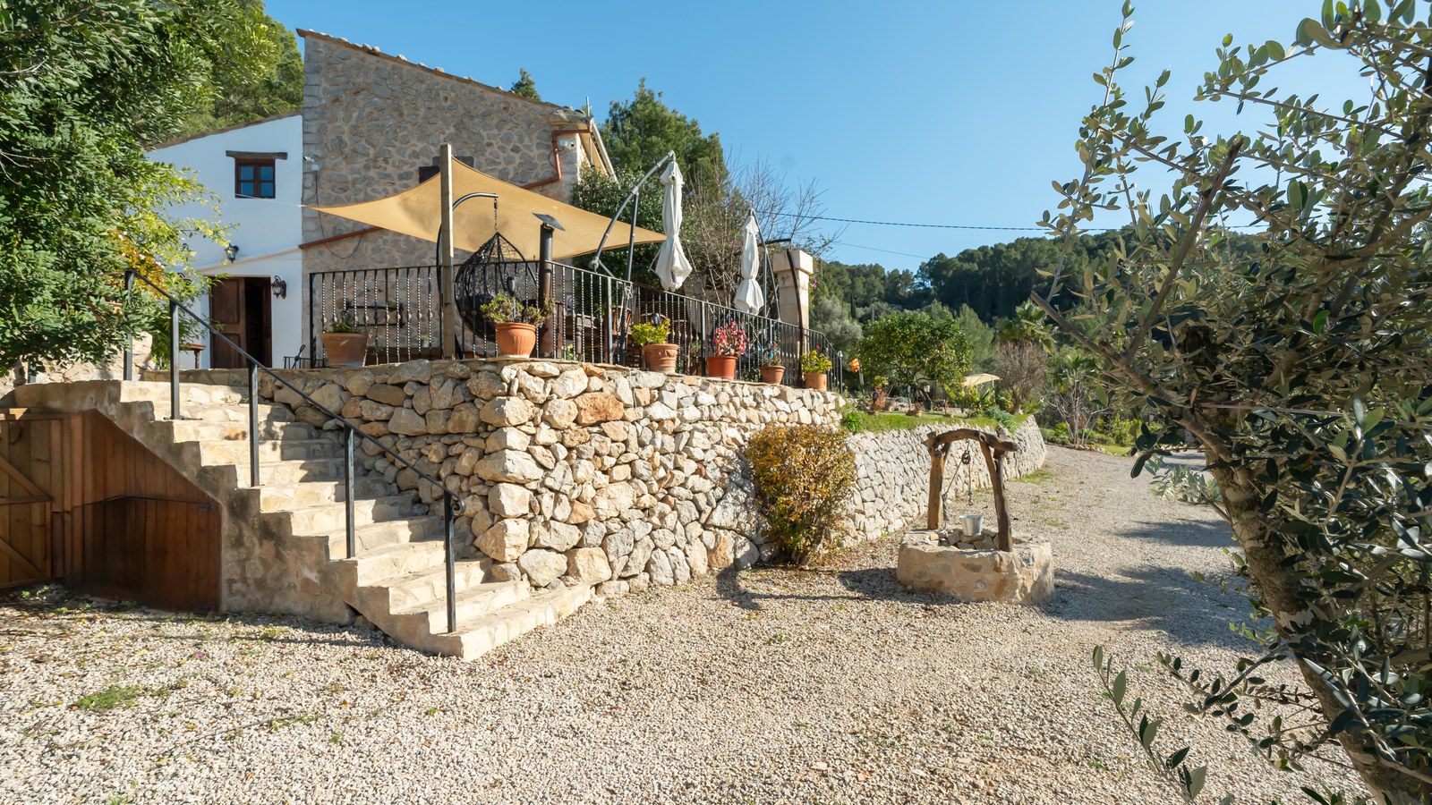 Terrasse surélevée en pierre avec voile d'ombrage, ferronnerie et puits traditionnel dans un paysage rural.