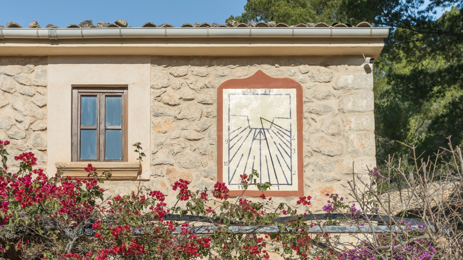 Cadran solaire artisanal sur une façade en pierre naturelle avec des bougainvilliers rouges sous le soleil.