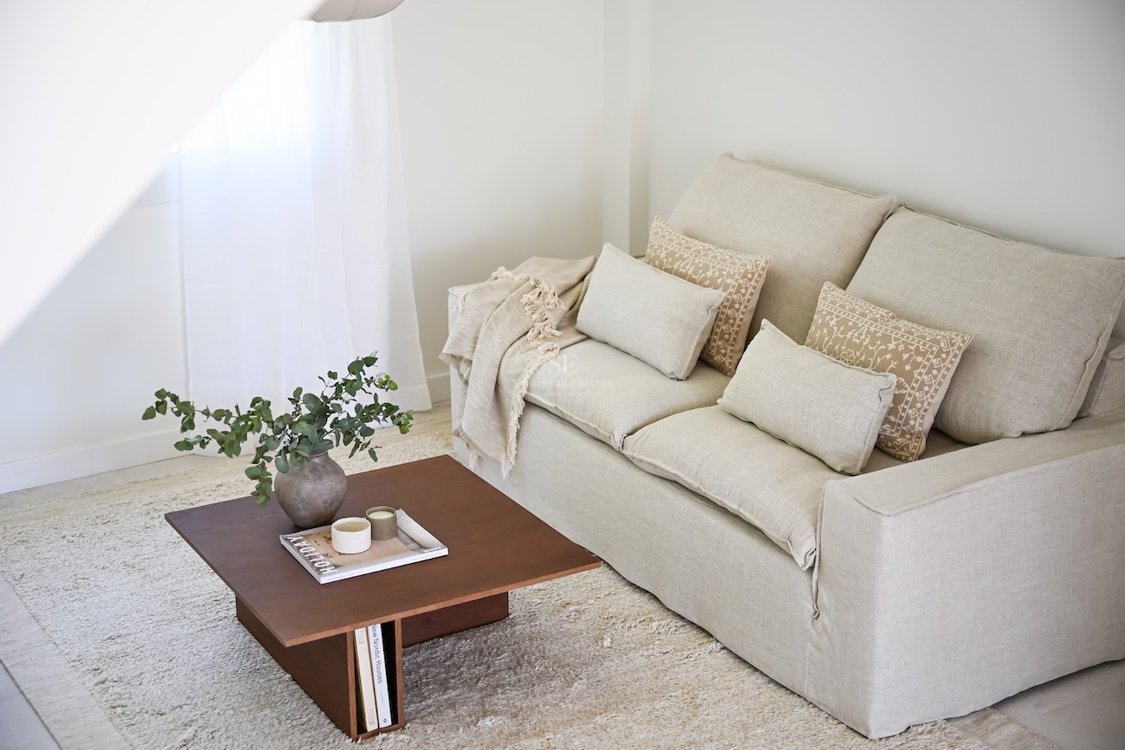 Salón minimalista con sofá de lino beige, mesa de centro de madera con eucalipto y alfombra de color crema texturizada.